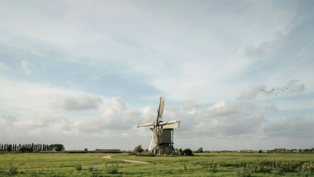 A traditional windmill standing tall in a lush green field under a cloudy blue sky with birds flying.