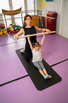 Physiotherapist holding wooden dowel and exercising with little girl indoors in the gym