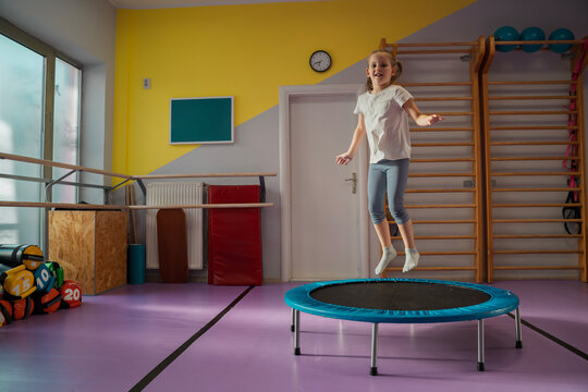 One cute little girl jumping on trampoline alone in the gym indoors, smiling and jumping while sun rays coming through the window 