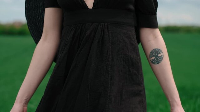 Close-up motion scene showing woman stepping through tall green grass wearing loose black dress, decorative mesh veil and tree forearm tattoo, with flowing fabric, soft overcast light and natural