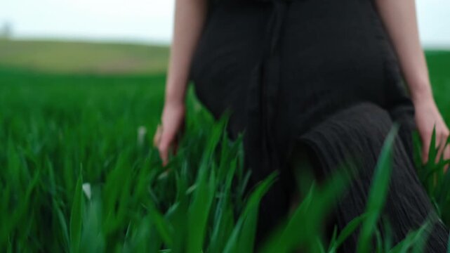 Low angle motion view of person taking small forward step through dense tall green grass, with long black textured skirt, visible hand movement, soft natural light, calm rural atmosphere, and
