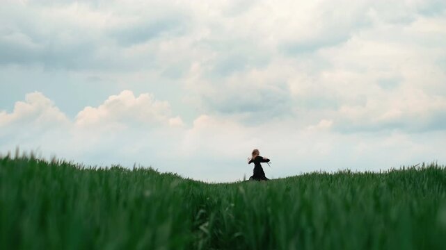 Young woman moves through quiet pose changes on grassy ridge in open field: seated with arms behind her, leaning slightly forward, standing upright, then returning to seated position while gazing