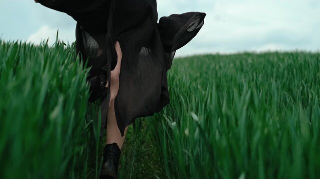 Close rear view shows lower body walking along narrow dirt path through dense tall green grass, with black skirt flowing, boots stepping forward, soft overcast light, and strong natural depth