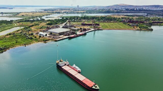A large cargo ship is anchored in the river near an industrial site. Green fields extend along the shore. In the distance, a city skyline can be seen under a clear sky.