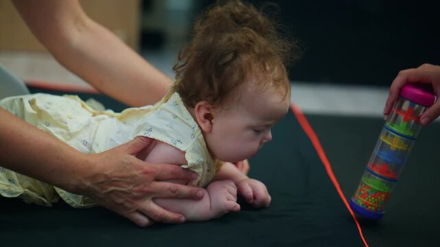 Close up of a baby reaching toward a colorful toy while being gently supported by a caregiver during mobility exercises