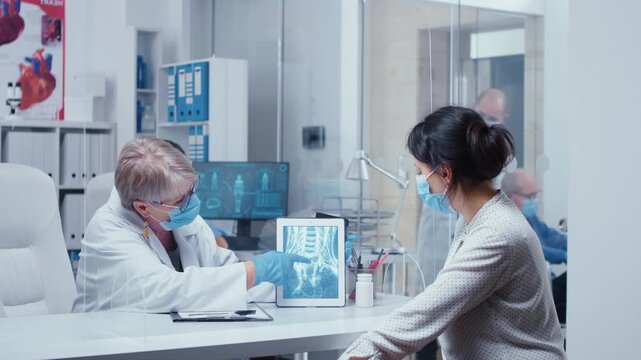Senior woman doctor shows an X ray on a tablet while wearing mask and gloves, consulting a patient through a plexiglass wall during protected clinical care.