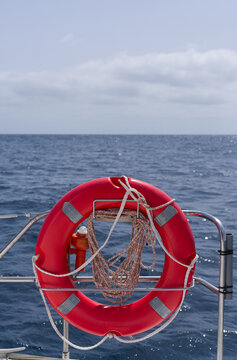 A bright red lifebuoy mounted on the stern of a sailboat with the open sea and blue sky in the background. Ideal for concepts of marine safety, sailing, travel, navigation, rescue, and adventure.
