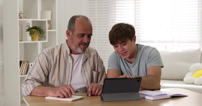 Caregiver. Teenager helping senior man learn how to use tablet at wooden table indoors
