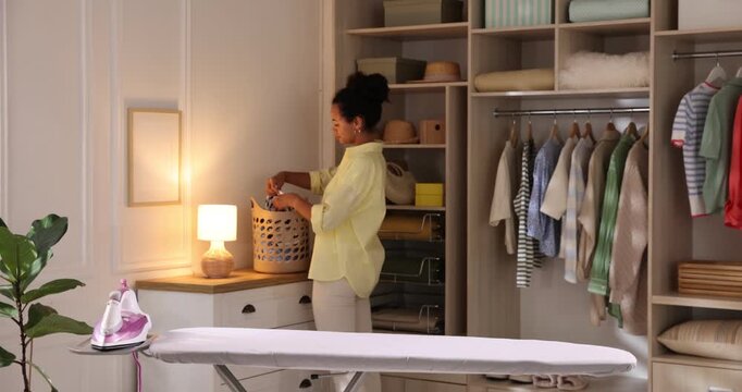 Woman putting laundry basket onto nightstand and ironing long sleeve shirt on board at home