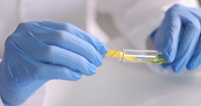Scientist holding PH test strip and glass tube with liquid at white table in laboratory, closeup