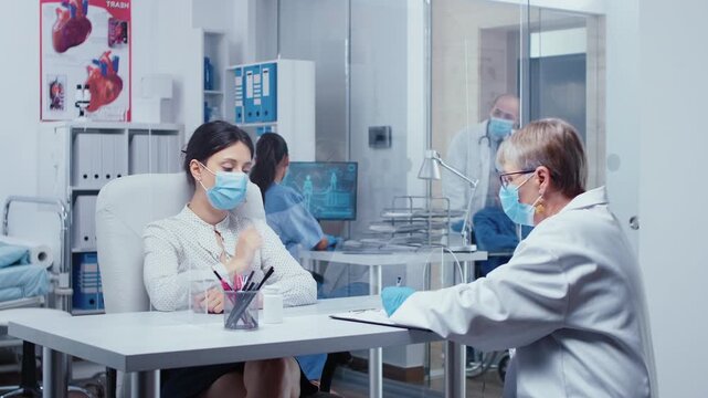 Doctor and patient wear masks while talking through a plexiglass wall during a protected medical consultation in a clinic office with clinical safety.