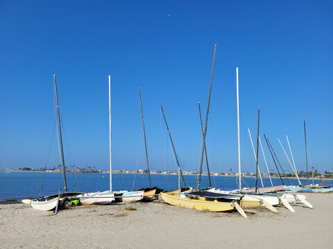 A line of catamarans and small sailboats parked on the sandy shore of Mission Bay under a clear blue sky in San Diego California