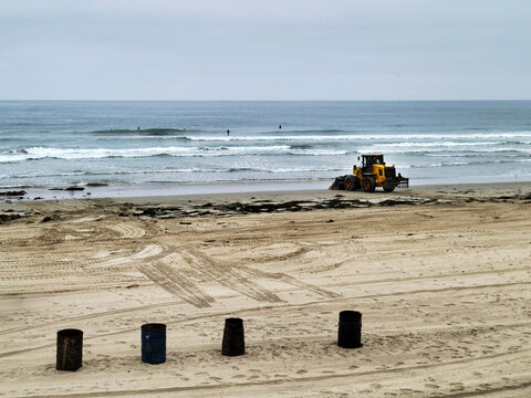 A yellow heavy tractor works on a sandy Pacific beach clearing seaweed near breaking ocean waves under an overcast sky in San Diego, California