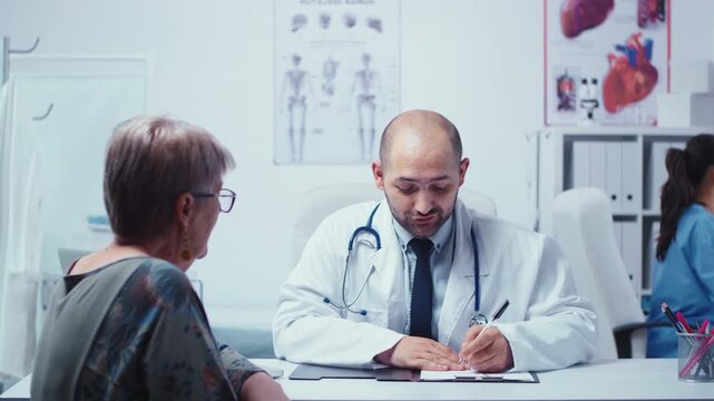 Male doctor interviews an elderly woman patient in his cabinet during a private clinic visit, gathering information for diagnosis and treatment.
