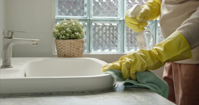 Close up of Asian Woman Hand Spraying Detergent and Wiping Bathroom Sink with Microfiber Cloth, Housekeeping Hygiene Routine in Soft Morning Sunlight