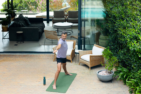 Mature man practicing standing yoga on green mat wearing purple T-shirt and black shorts on patio