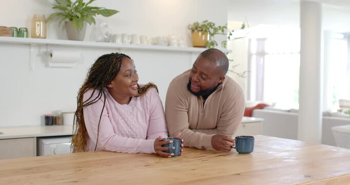 African couple sitting at wooden table, woman in pink talking causing man leaning with blue mugs