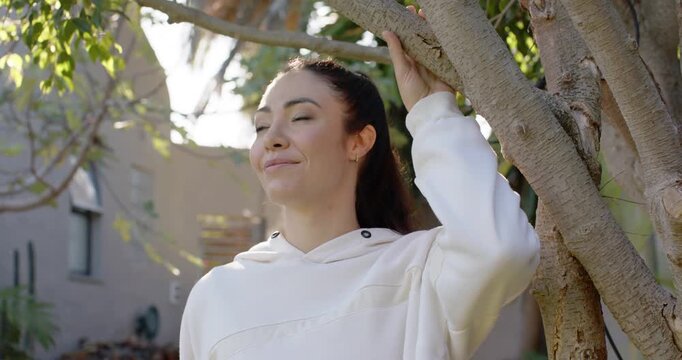Woman standing in yard holding branch in light hoodie, turning face and noticing sunlight in leaves
