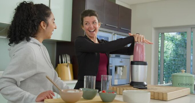 Diverse women watching in kitchen while right woman pressing blender cup preparing pink smoothie