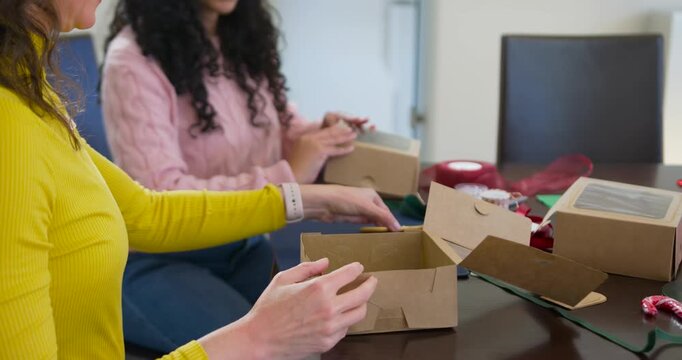 Two adult women seeing flat boxes, folding and assembling gift boxes at table, preparing gifts