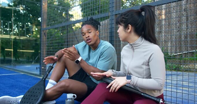 Diverse training partners sitting on blue court edge after play, discussing strategy with paddles