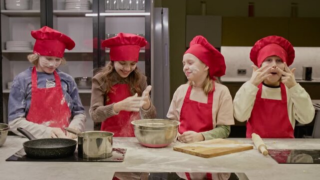 Caucasian kids wearing red chef hats putting flour on faces and laughing during baking class. Ideal for promoting culinary schools or children cooking programs