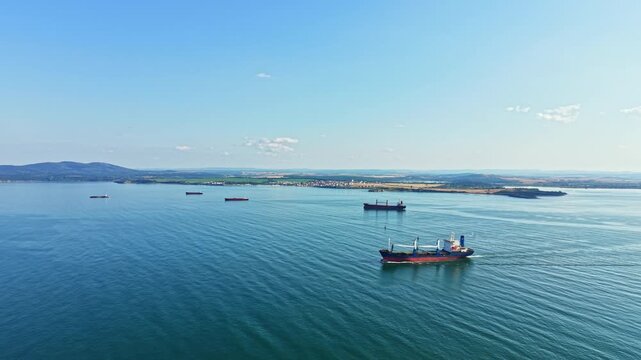 Several ships are sailing in a bay while a small town sits on the shore and hills rise in the background under a clear blue sky during a sunny afternoon.
