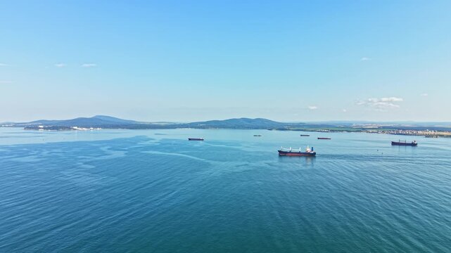 Several large ships move slowly on the water near a coastline. Hills are visible in the background, and the sky is clear. It is daytime and the sun is shining.