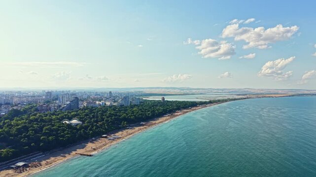People walk along the beach. Buildings stand near the shore. The water reflects sunlight. Trees line the coast. The sky has white clouds.