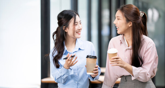 Two young Asian businesswomen chatting happily during coffee break in office.
