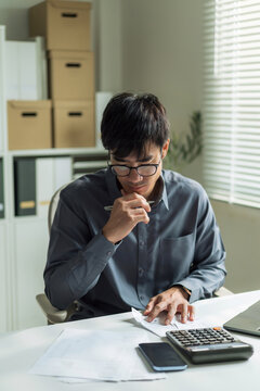 Stressed businessman wearing glasses holding chin while checking financial bills and using calculator at office desk.