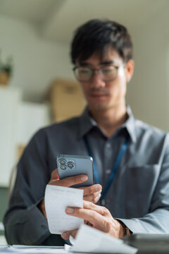 Vertical shot of young businessman using smartphone to check or scan receipt bill for financial tracking at desk.