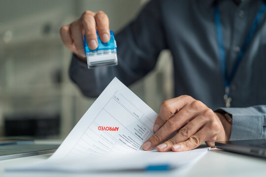 Close-up of office worker hand stamping red approved word onto business document contract.