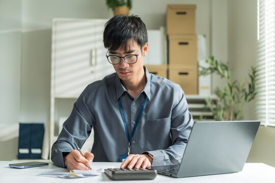 Focused young businessman wearing glasses writing notes and calculating documents at office desk.