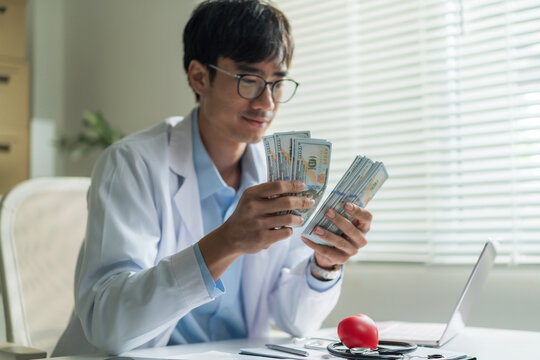 Young doctor wearing glasses smiling happily while counting stack of US dollar banknotes at clinic desk.