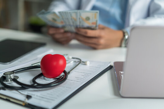 Close-up of red heart model and stethoscope on clipboard with doctor counting dollar bills blurred in background.