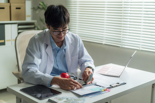 Young doctor in white coat counting US dollar banknotes at desk with heart model and stethoscope in clinic.