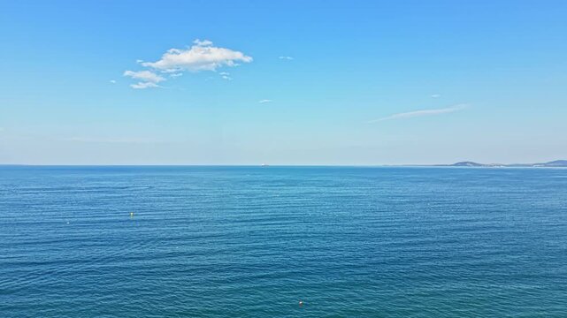 The scene shows a vast blue ocean stretching far into the distance under a clear sky with some clouds. Gentle waves move across the water with small boats visible on the horizon.