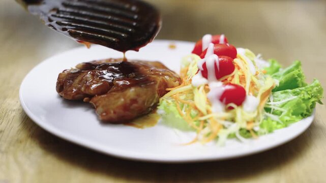 Juicy grilled meat steak being glazed with thick savory sauce. Served with a fresh side salad of shredded vegetables and cherry tomatoes with white dressing on a white plate over a wooden surface.