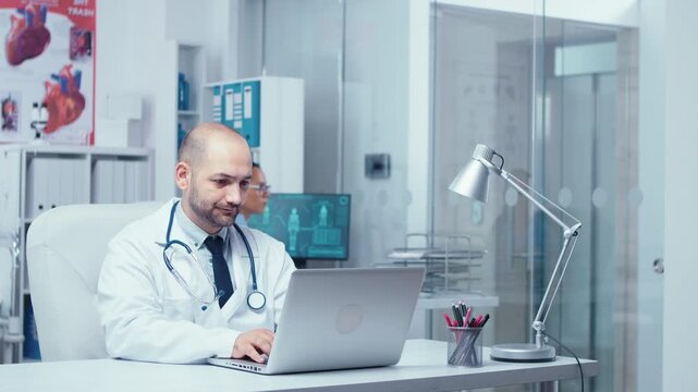Doctor types an email from a private clinic office while a nurse checks X rays and patients walk through a busy glass walled hallway.