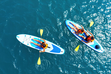 Fototapeta premium Couple paddle boards on clear blue water. People enjoy summer day on sup boards wearing life vests. They relax together during vacation on calm ocean.