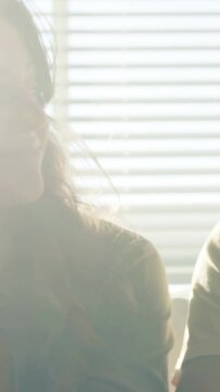 Vertical video: Smiling woman turning during chat on couch at home, sunlit blinds, partner still