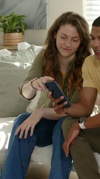 Vertical video: Leaning, woman in olive top on couch taking blue phone from man to view screen