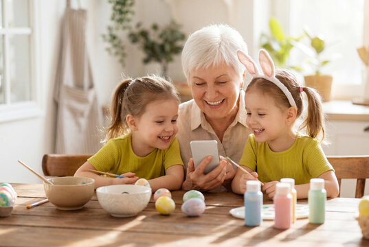 Grandmother and twin granddaughters looking at a smartphone while painting easter eggs at home