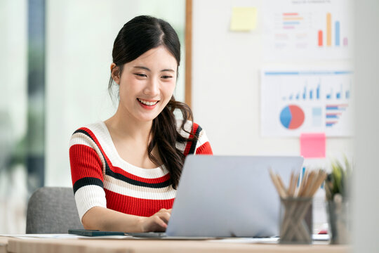 Happy young Asian businesswoman smiling while working on a laptop in modern office.