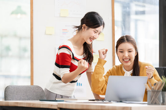 Two Cheerful Asian Businesswomen Celebrating Success While Working on Laptop in Office