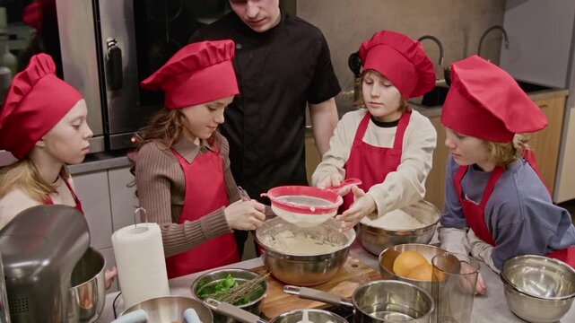 Caucasian male chef instructing group of Caucasian children during culinary workshop, kids whisking and sifting ingredients. Ideal for promoting children cooking classes or kitchenware brands