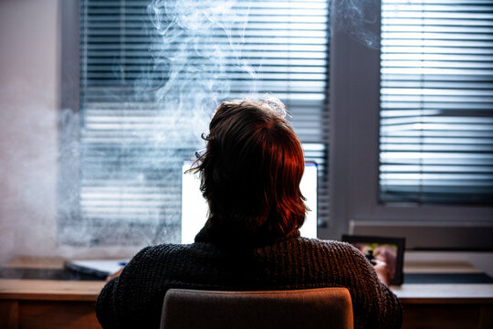 man seen from behind sitting by a bright window with soft smoke rising in a moody contemplative interior scene.
