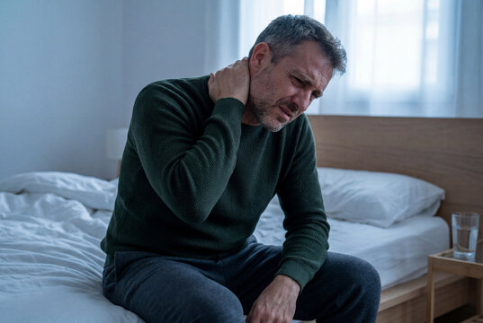 Middle aged man touching sore neck sitting on edge of bed