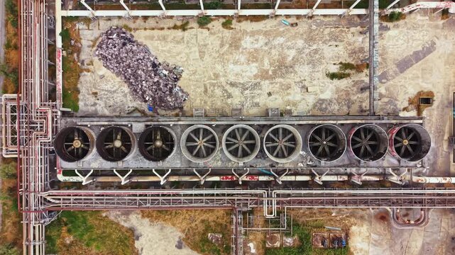 Aerial view of an industrial site featuring several cooling towers arranged in a row. Pipes connect the towers and debris piles are visible on the ground.